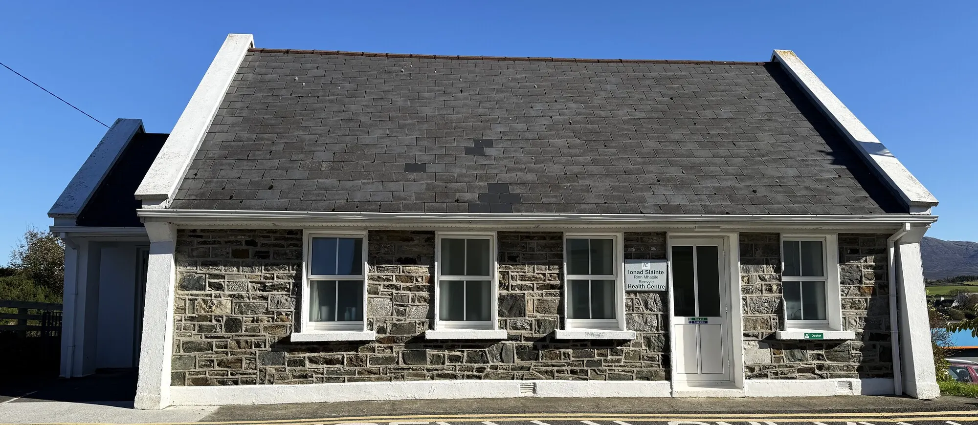 Renvyle Health Centre in Tully: a stone-fronted single-storey building with white gables, under a clear blue sky.