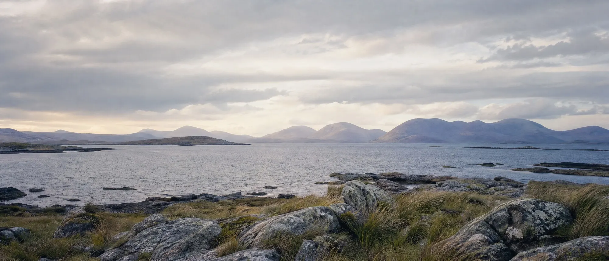 A calm Atlantic shoreline with stone, sea grass and distant mountains in North Connemara.